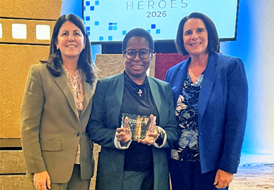 Three women stand smiling, with the center woman holding a clear award, in front of a "HEROES 2026" backdrop at an indoor event.