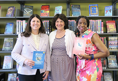Three women standing in a library, smiling and holding books titled "White Coats" in front of bookshelves filled with various books.