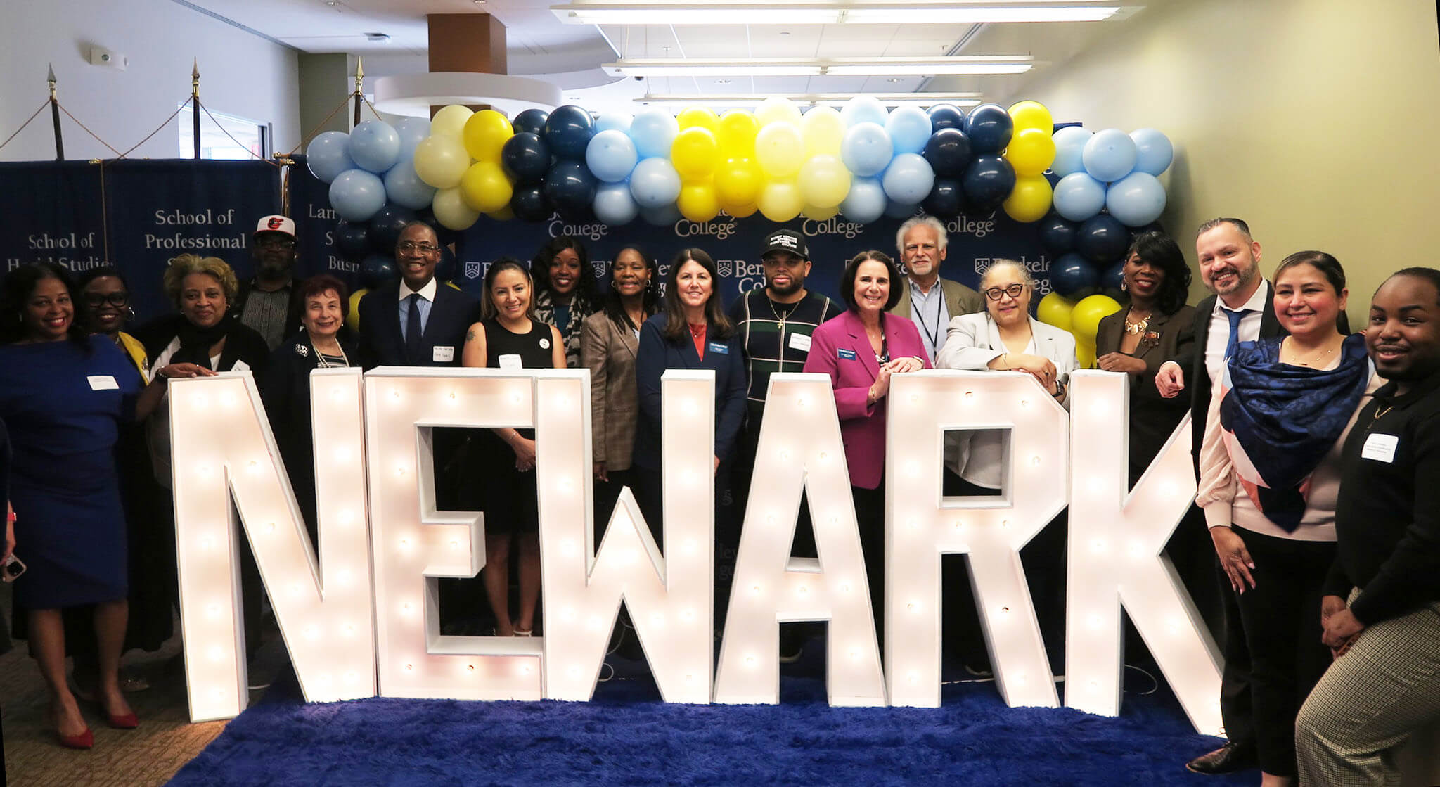 Group of people posing behind large illuminated letters spelling "NEWARK" with blue, white, and yellow balloons in the background.