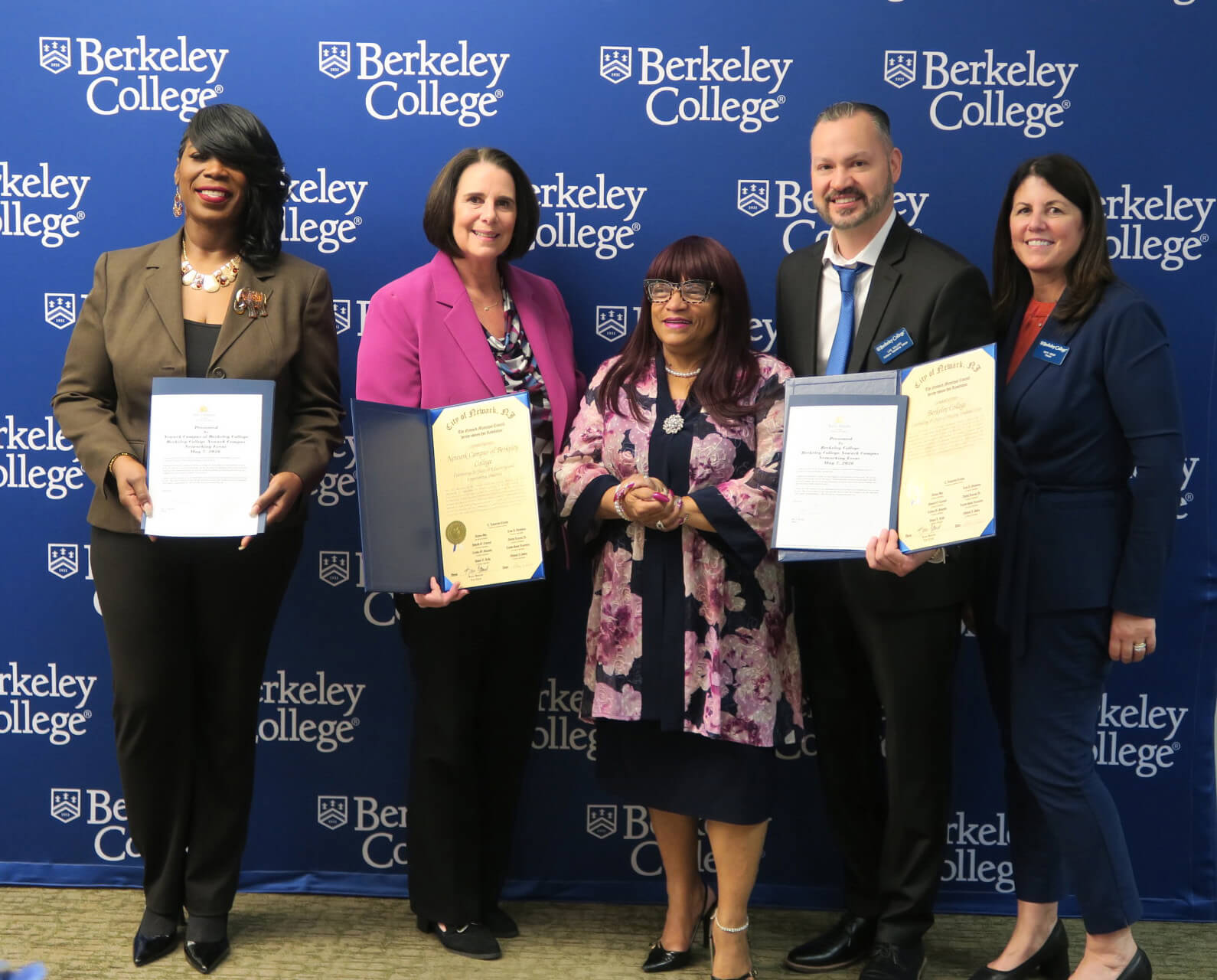 Five people stand in front of a Berkeley College backdrop, three holding certificates, dressed in formal business attire.