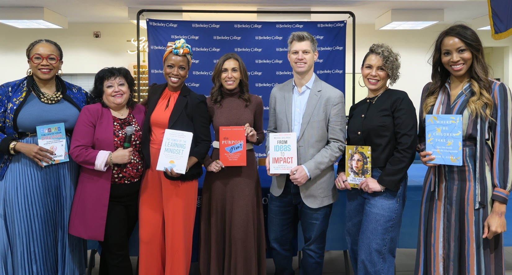 Seven diverse people stand smiling in a row, and six of them hold books they authored.