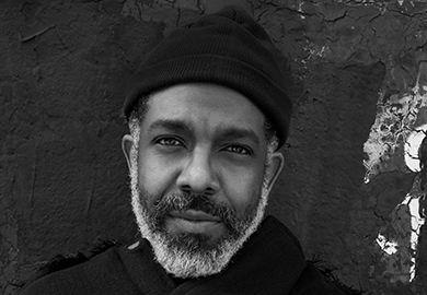 Black and white close-up portrait of a bearded man wearing a beanie, standing against a textured, dark wall.