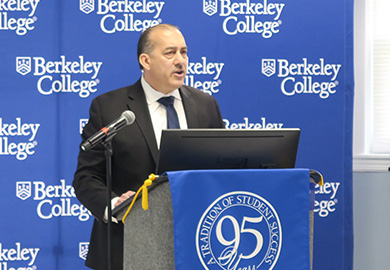 Person in suit standing at a podium with a microphone, giving a speech at Berkeley College event with blue branded backdrop.