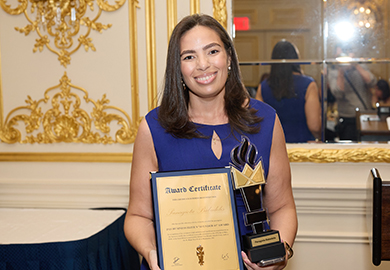 Person in a blue dress holding an award certificate and a trophy featuring a gold crown design in an ornate room.
