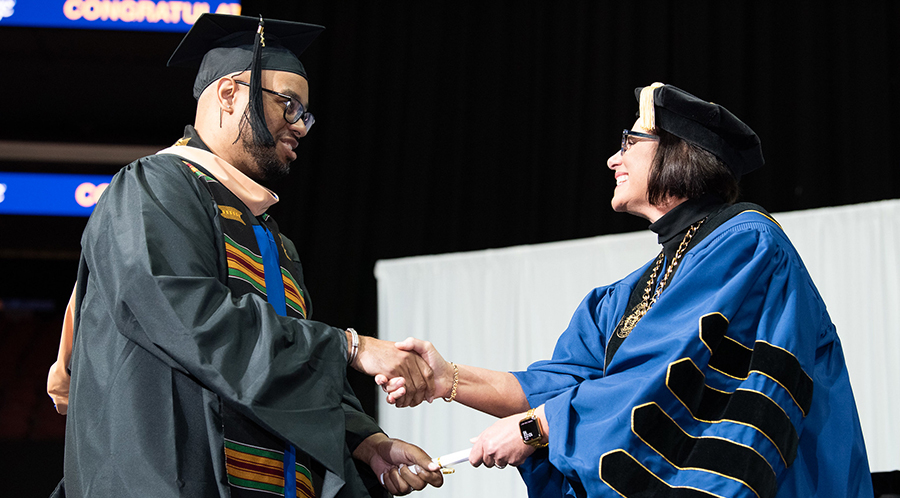 a male student in cap and gown accepts a diploma from a woman in a commencement robe