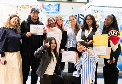 Group of people posing indoors, some holding certificates, with festive decorations and a television screen in the background.