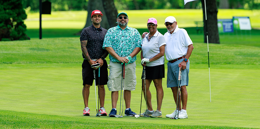 Four diverse people posing with golf clubs on a lush green golf course near the hole flag on a sunny day.