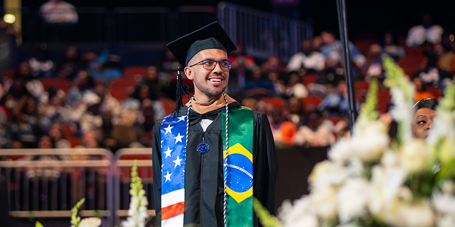 Graduate wearing a cap and gown with American and Brazilian flag stoles, standing at a ceremony with blurred audience in the background.