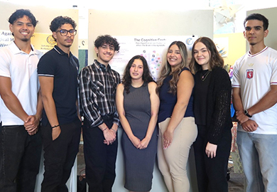 Group of six young adults standing together indoors, smiling in front of a presentation poster.