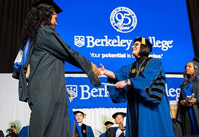 Graduate in cap and gown shakes hands with college official in robe at Berkeley College 95th anniversary ceremony.