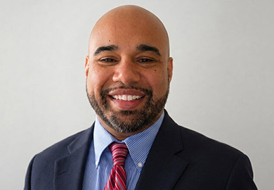 Smiling man with a beard wearing a dark suit, blue shirt, and red striped tie against a light background.