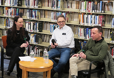 Three people holding microphones sit in a library setting, engaged in conversation with bookshelves behind them.