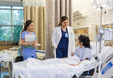 Two healthcare professionals talk with a patient lying in a hospital bed in a bright, well-equipped hospital room.