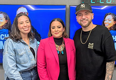 Three people smile at the camera in front of blue screens: two women and a man wearing a Yankees cap and black shirt.
