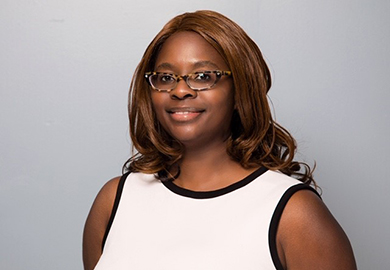 Portrait of a smiling woman with glasses and shoulder-length brown hair, wearing a sleeveless white top against a gray background.