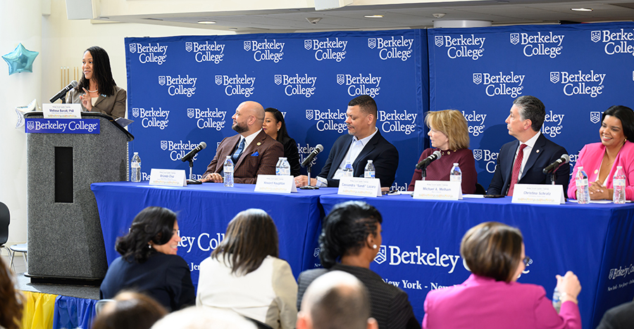 a panel of people sit on a stage with a woman at the podium