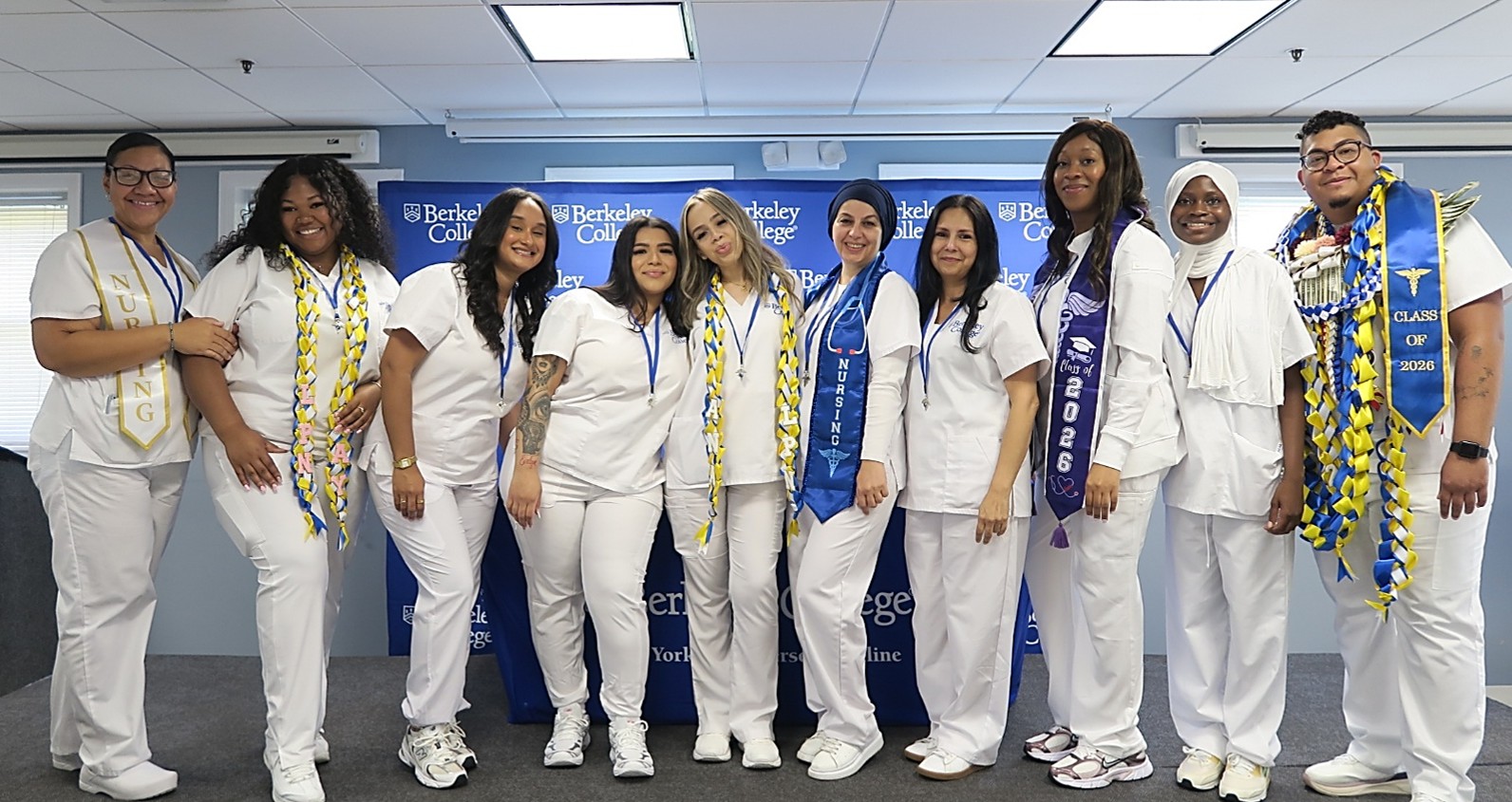 A group of diverse students in white scrubs stand in a line and smile at the camera.