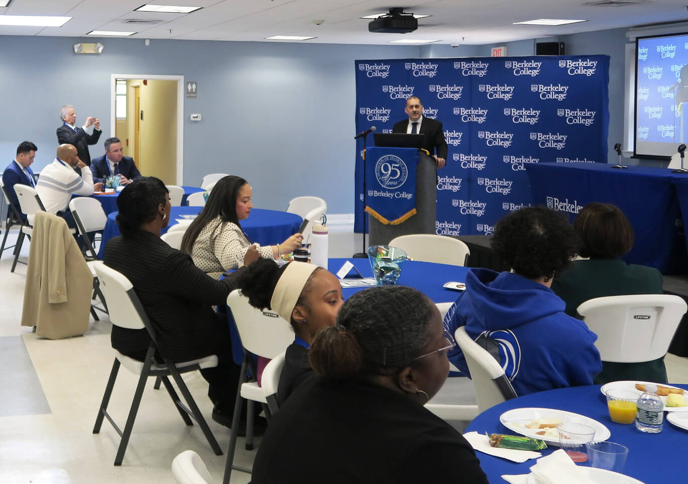 A speaker at podium addressing a seated audience in a conference room with Berkeley College banners.