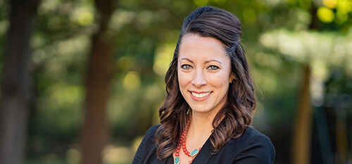 A woman with crossed arms poses confidently in front of a backdrop of vibrant trees