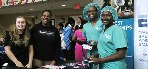 our women stand beside a table displaying a sign. Two women wear nursing scrubs showcasing a professional healthcare environment, as they talk to the other two women in casual clothes, who promote their company as a potential employer.